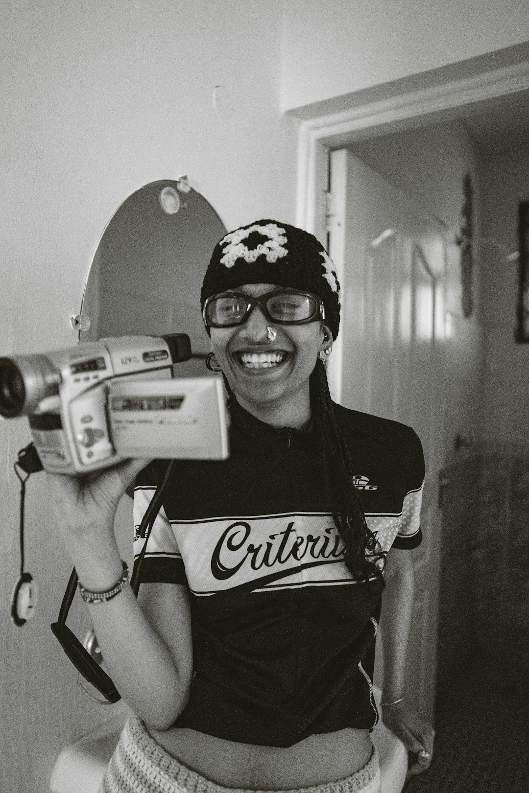 Smiling woman holds old camcorder, wearing beanie and glasses.